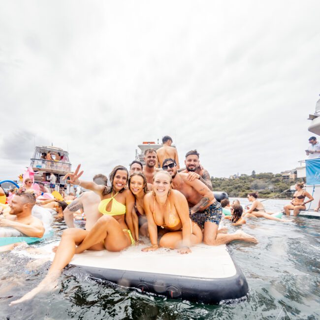 A group of five people in swimwear pose cheerfully on an inflatable raft in the water. In the background, other people are enjoying the water, with boats and a cloudy sky visible. The photo has a fun, summer party vibe reminiscent of Boat Parties Sydney by The Yacht Social Club. "No swipes" is written in the bottom left corner.