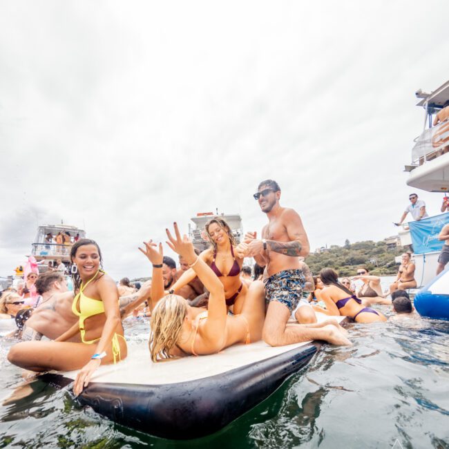 A group of people in swimwear are enjoying themselves on a boat and in the water. They are lounging on a large floating air mattress, with some smiling and posing for the camera. The lively scene by The Yacht Social Club Sydney Boat Hire is set against a cloudy sky and calm water.