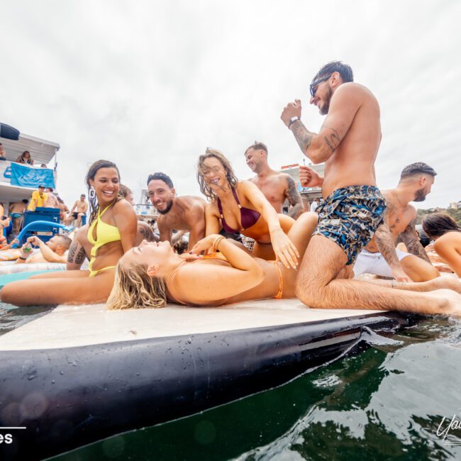 A group of people in swimsuits are enjoying themselves at a waterfront party. Some are lounging on an inflatable raft in the water, while others smile and laugh around them. The atmosphere is lively and social with a party boat from The Yacht Social Club Sydney Boat Hire visible in the background.