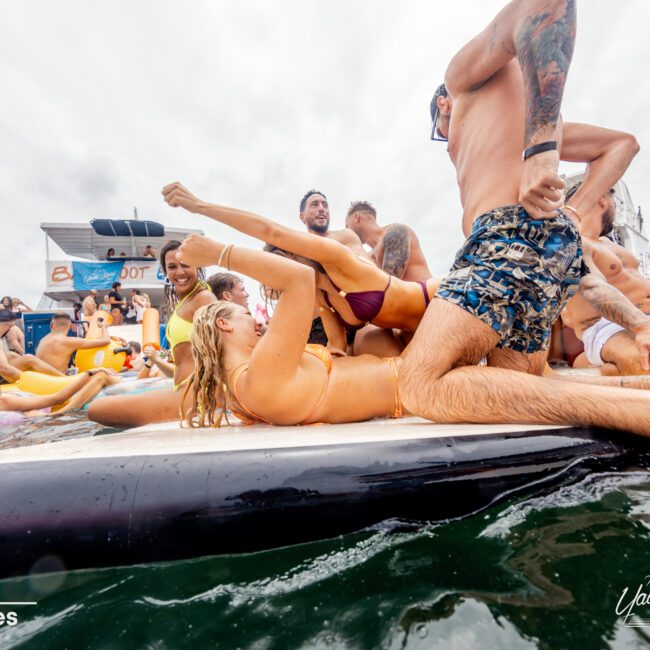 A group of people in swimwear are enjoying themselves on a floating platform or inflatable in the water, part of The Yacht Social Club Event Boat Charters. They appear to be at a crowded, festive event with boats and other inflatables in the background. The atmosphere is lively and spirited.