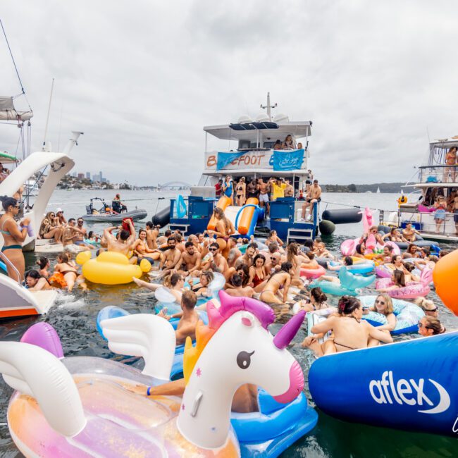 People are having fun at a boat party, floating on colorful inflatable rafts and engaging in lively activities on the water. The scene includes various boats parked closely together, with people socializing, swimming, and enjoying the festive atmosphere of The Yacht Social Club Sydney Boat Hire.