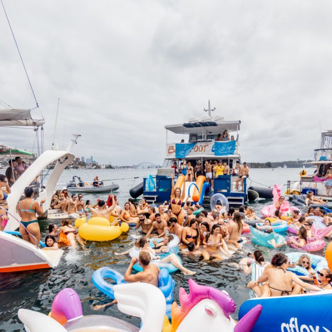 A lively boat party in Sydney features numerous colorful inflatables, including a unicorn and a swan, with many people enjoying the water around a large blue and white yacht. The event is hosted by The Yacht Social Club against a backdrop of a cloudy sky and distant cityscape.