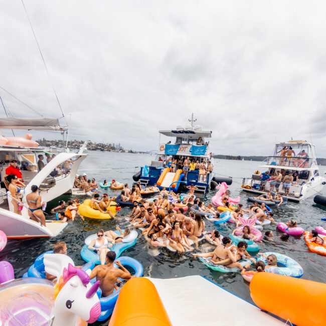 A large group of people enjoying a party on the water with various pool floats, including a unicorn. Several festively decorated boats dock nearby, embodying the vibrant spirit of Boat Parties Sydney with The Yacht Social Club. The cloudy sky and distant shore add to the lively, social atmosphere.