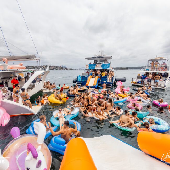 A lively party scene unfolds on the water, with numerous people enjoying colorful inflatable floats and pool toys. Surrounding them are several yachts and boats, some equipped with slides. The weather is overcast, but the atmosphere from The Yacht Social Club Sydney Boat Hire remains festive and energetic.