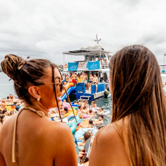 A lively scene at *The Yacht Social Club* event on the water, featuring numerous people in swimsuits enjoying the festivities. Two women in the foreground look towards the main activity, with boats and a crowd of partygoers on inflatables visible in the background.