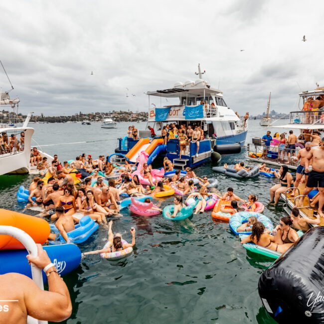 A bustling scene of people on colorful inflatable floats surrounds a few anchored boats on a cloudy day. The crowd appears to be enjoying a lively social event organized by The Yacht Social Club Sydney Boat Hire, with some people onboard the boats and others in the water, while seagulls fly overhead.