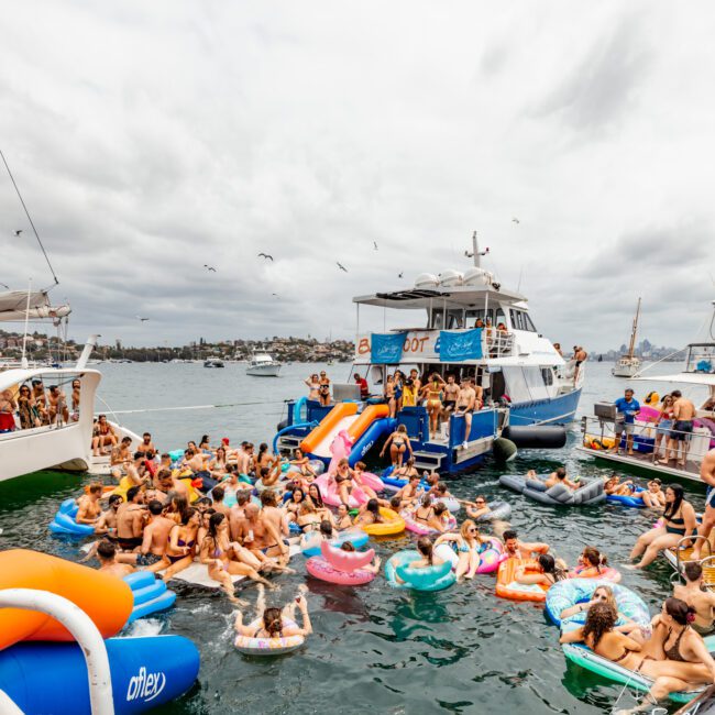 A large gathering of people are enjoying a boat party on a cloudy day. Many are on colorful inflatable floats in the water, surrounded by boats. The scene is lively with people swimming, socializing, and enjoying the festive atmosphere at The Yacht Social Club Event Boat Charters.