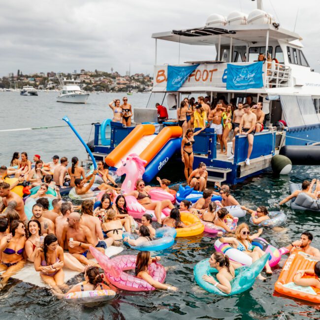 Many people are enjoying a pool party on inflatable floats in the water next to a large yacht with a slide facilitating entry. The sky is cloudy, and more boats are in the distance. A banner "Barefoot" is visible on the yacht, part of The Yacht Social Club Sydney Boat Hire.