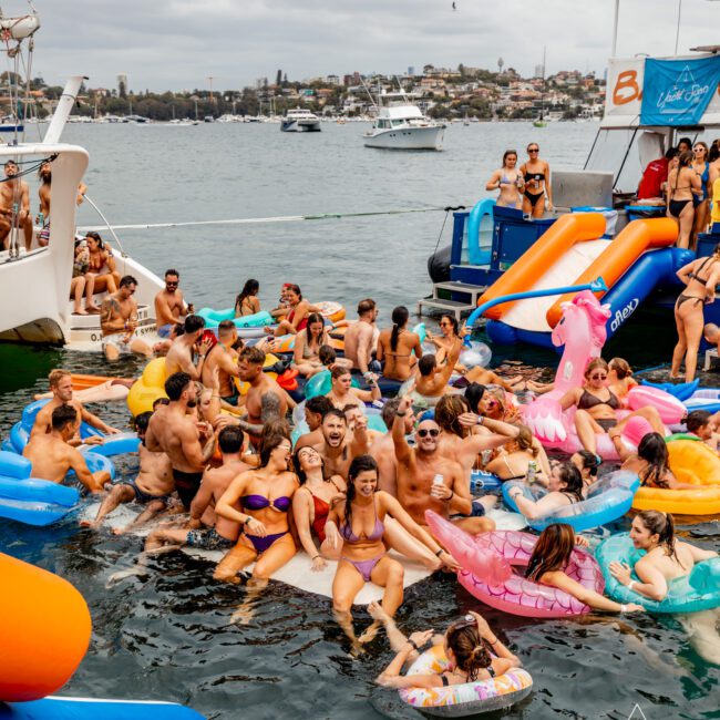 A lively group of people having fun in the water with colorful inflatable floats, including flamingos and unicorns, near several docked boats. The festive atmosphere showcases participants wearing swimsuits against a cityscape backdrop, all part of Boat Parties Sydney The Yacht Social Club event.