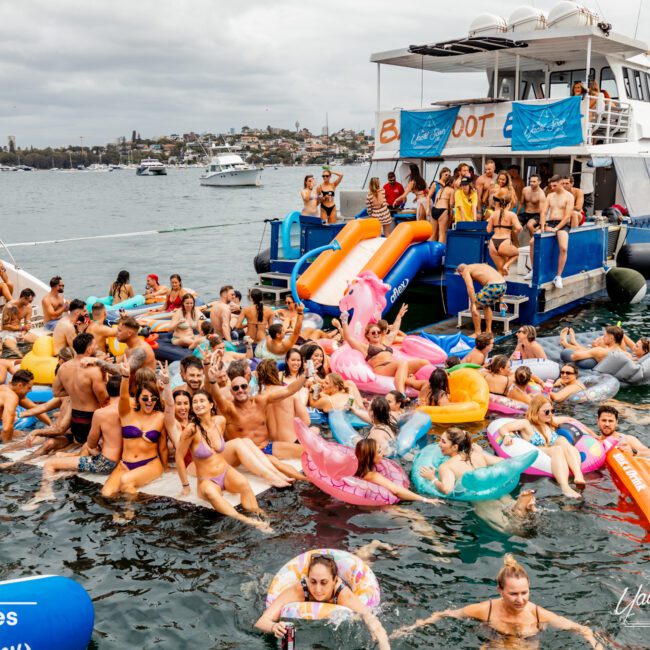 A lively group of people in swimsuits are enjoying a party on a boat and in the water, surrounded by colorful inflatable floats. Another boat is docked nearby, and the background shows a bay with other boats and a cloudy sky. It's all part of The Yacht Social Club Event Boat Charters experience.