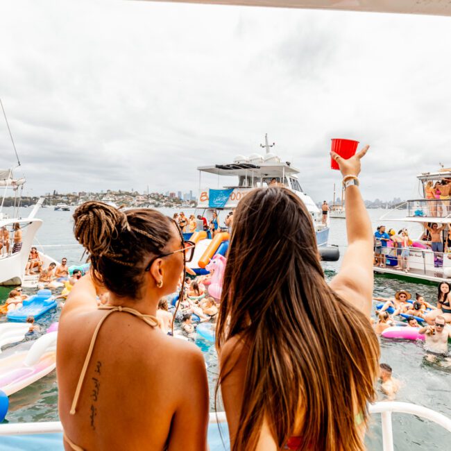 A large group of people is enjoying a lively boat party hosted by The Yacht Social Club. Two women in the foreground, one raising a red cup, face a crowd of people on boats and inflatables in the water. The scene is festive with various boats anchored close together under a cloudy sky.