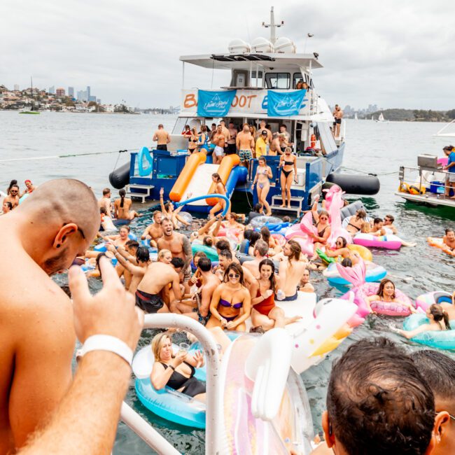 A lively boat party with numerous people enjoying themselves on a docked yacht and colorful inflatables in the water. Most are in swimwear, with some holding drinks. The yacht features a slide, and cityscape buildings are visible in the backdrop. Text reads "The Yacht Social Club Event Boat Charters.