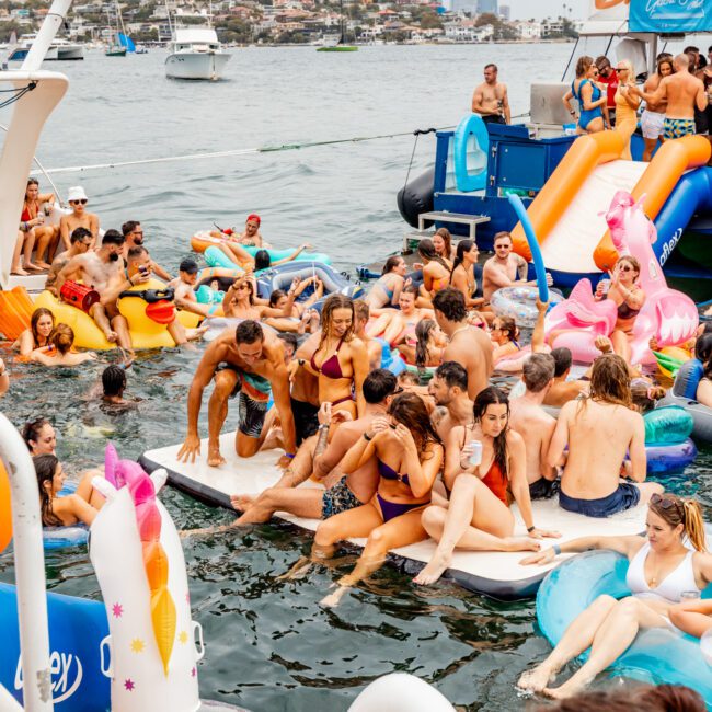 A vibrant scene of a floating party on a body of water. People are lounging on inflatables, floating mats, and boats. Various colorful floats shaped like swans and flamingos add to the festive atmosphere. The Yacht Social Club Sydney Boat Hire in the background features city buildings and boats.