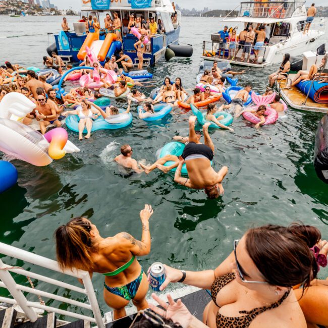 People are enjoying a lively boat party hosted by The Yacht Social Club. Many are swimming or floating on inflatable rafts near two large boats. Some are jumping into the water from a platform, while others relax on board. The scene is festive with the crowd engaged in different activities around Sydney Harbour.