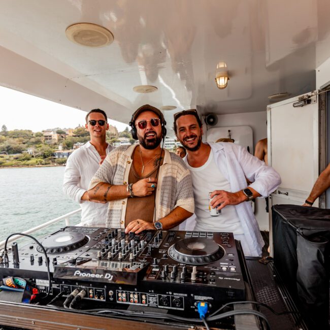 Three men smile while posing on a boat with DJ equipment in front of them during a lively Boat Parties Sydney The Yacht Social Club event. The man in the middle sports sunglasses and headphones around his neck. In the background, water and greenery can be seen, with a life preserver mounted on the wall to the right.