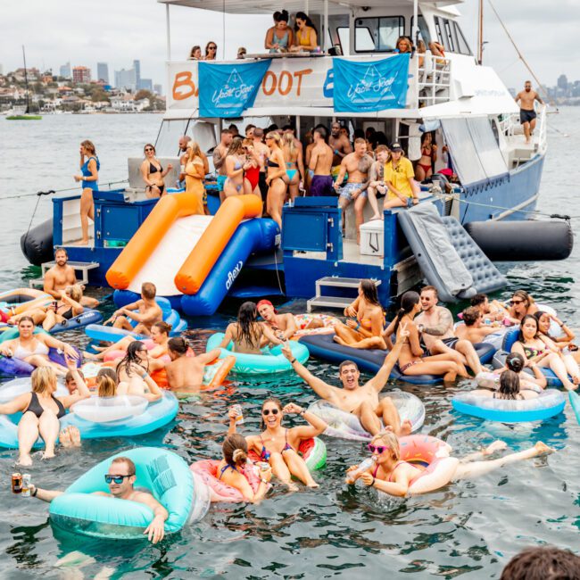 A large group of people enjoying a boat party: many are on inflatable floats in the water, while others are on the yacht with an orange slide. They appear to be having fun under cloudy skies. The yacht displays a banner "The Yacht Social Club," part of Sydney Harbour Boat Hire. Logos for branding are visible.