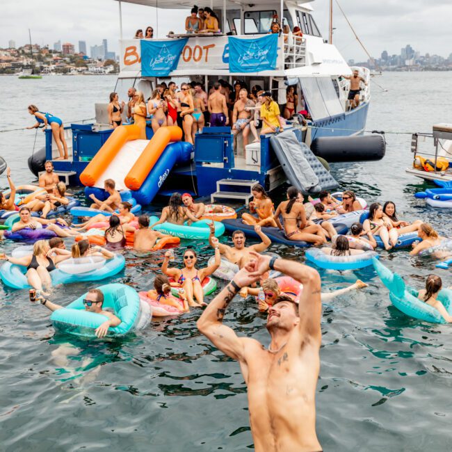 A lively boat party scene with numerous people swimming and floating on inflatable rings in the water. Several individuals are gathered on a large boat with a slide, part of The Yacht Social Club Sydney Boat Hire. The boat is anchored in a bay with a cityscape visible in the background under an overcast sky.