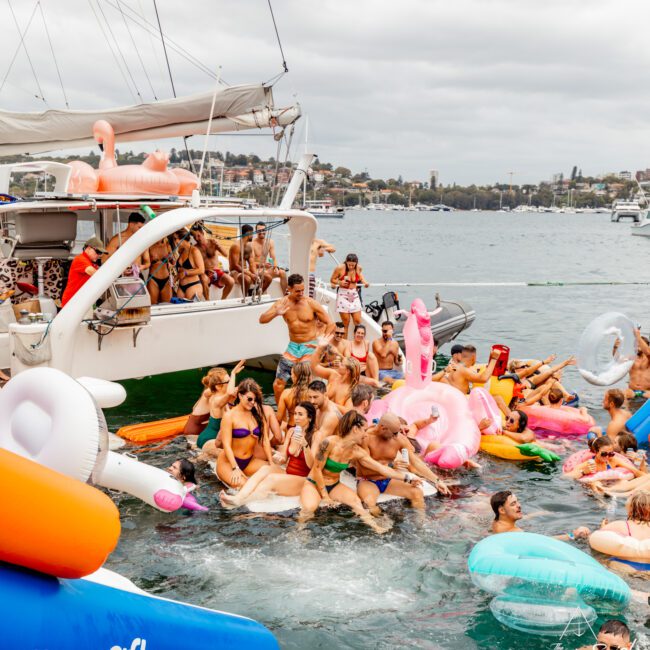 A large group of people enjoying a yacht party on the water. Various individuals are in swimsuits, many floating on inflatable pool toys such as flamingos and unicorns. The Yacht Social Club Event Boat Charters offers this unforgettable experience near a scenic coastline with buildings in the background.