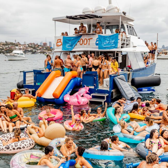 A large group of people in swimwear enjoy a sunny boat party hosted by The Yacht Social Club on Sydney Harbour. They are on and around a luxury yacht decorated with inflatable toys, like flamingos and unicorns. Several guests slide off colorful slides into the water, with other boats seen in the background.