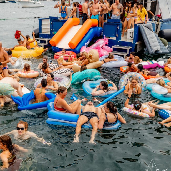 A lively scene of people enjoying a party in and around a boat, with many floating on colorful inflatable rafts in the water. Some are on the boat, socializing and using the water slides. The setting appears festive with lots of laughter and merriment, typical of a Sydney Harbour Boat Hire The Yacht Social Club event.