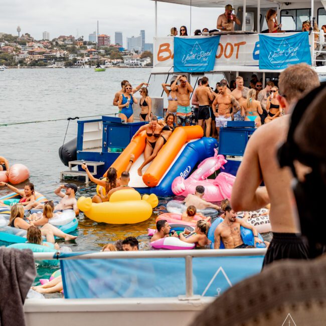 A large group of people in swimwear are enjoying a boat party on the water. Inflatable floats are scattered around. Two large blue and orange slides are seen on the boat. Buildings and a cityscape are visible in the background under a cloudy sky, hosted by The Yacht Social Club Sydney Boat Hire.