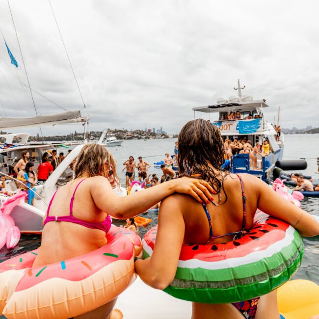 Two people in festive inflatable rings, one shaped like a watermelon and the other like a donut, stand arm in arm, looking towards a crowd of partygoers on boats and inflatables. The lively scene of Boat Parties Sydney The Yacht Social Club has everyone enjoying the water under a cloudy sky.