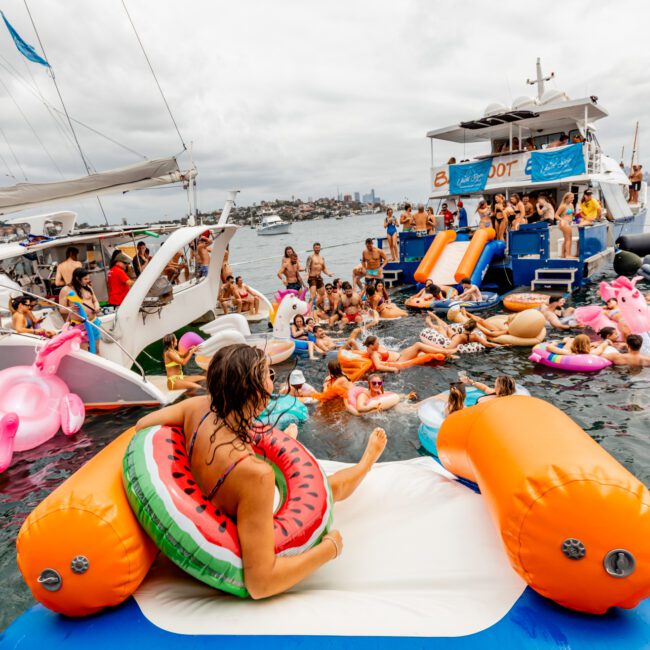 A lively scene on a waterway features people on various inflatables, including a watermelon float, between two boats. Attendees from The Yacht Social Club Sydney Boat Hire are socializing and enjoying themselves in the water, with the backdrop of a cloudy sky and distant shoreline.