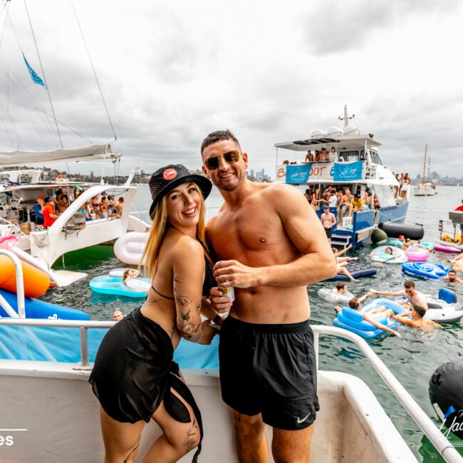 A smiling woman and man in swimwear pose on the deck of a boat, with other people on swim floats and additional boats in the background. The lively atmosphere at The Yacht Social Club is highlighted by the crowd and numerous inflatables. The sky is overcast. Text reads "No swipes.