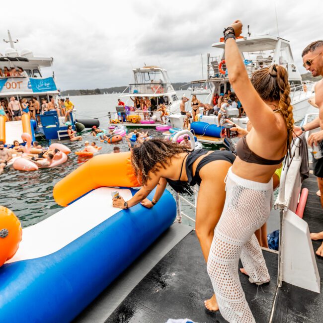 A lively scene at The Yacht Social Club Sydney Boat Hire party with people both on the boat and in the water. Colorful inflatables fill the water, and individuals in swimsuits are dancing and enjoying themselves. Several boats are visible in the background, under a cloudy sky.