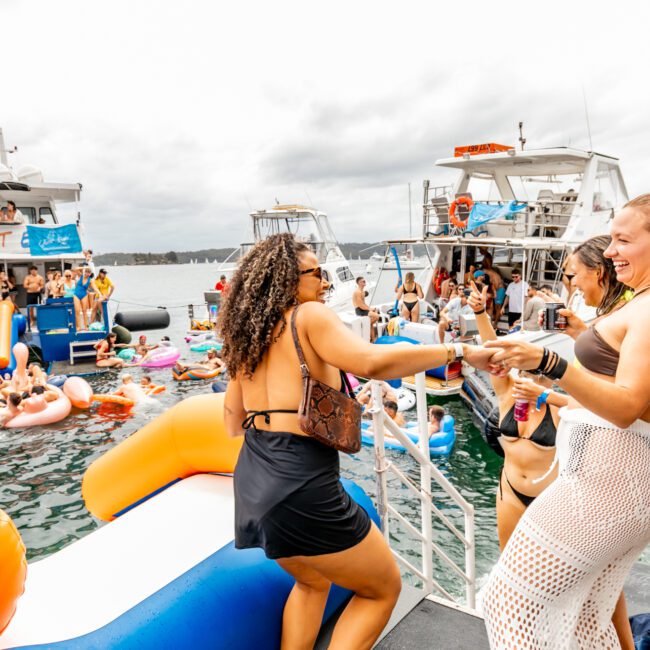 A lively scene of people enjoying a party on boats and in the water. Two women are laughing and dancing on a boat's edge while others swim and float on inflatables nearby. The backdrop features cloudy skies and more partygoers on adjacent boats, all part of The Yacht Social Club Sydney Boat Hire event.