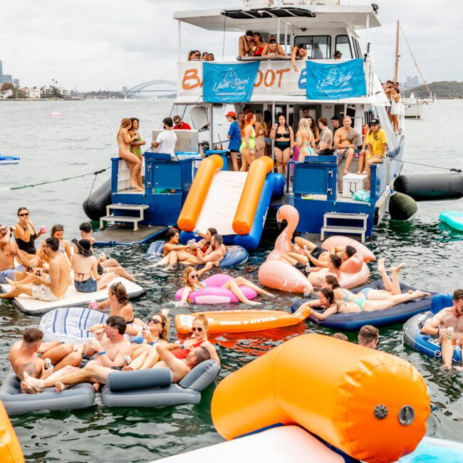 A vibrant boat party scene with numerous people enjoying themselves on a large yacht and surrounding floating inflatables in the water. The Yacht Social Club Event Boat Charters hosts the event, featuring fun slides and colorful floaties where everyone is relaxing. A banner reads "Boat Party.