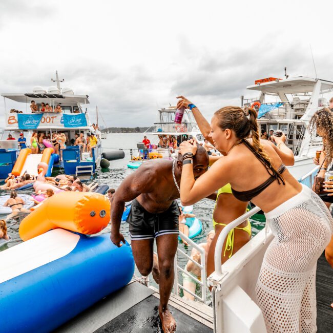 A lively scene on a yacht with people enjoying a party. In the foreground, a woman in a bikini and mesh cover-up playfully helps a man onto the boat. The background features more partygoers on inflatables, slides, and floating platforms, showcasing Boat Parties Sydney The Yacht Social Club.