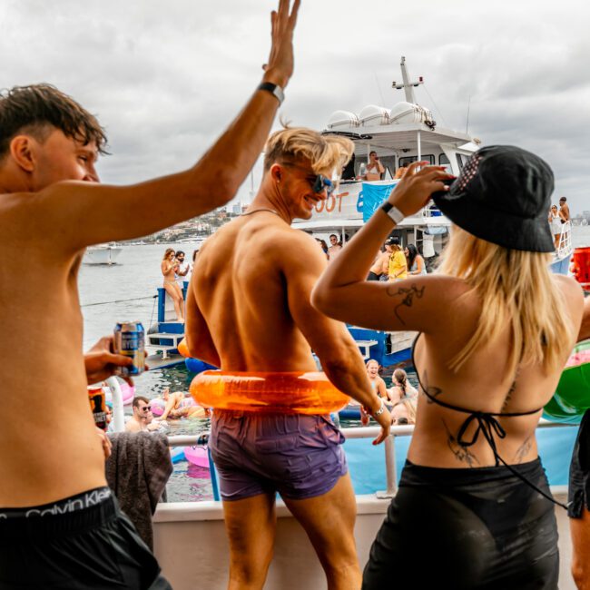 A group of people in swimsuits are on a boat, dancing and enjoying a party. Some are wearing inflatable tubes, and one person is holding a water gun. The scene is lively, with other boats and a cityscape in the background. It's a cloudy day, perfect for Boat Parties Sydney with The Yacht Social Club.