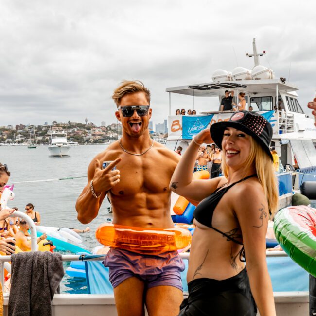 A group of people enjoying a lively boat party. In the foreground, a shirtless man and a woman in a black bikini are dancing and posing for the camera, both wearing sunglasses. Other partygoers relax and socialize behind them on the luxury yacht from Sydney Harbour Boat Hire against a backdrop of boats and a cloudy sky.