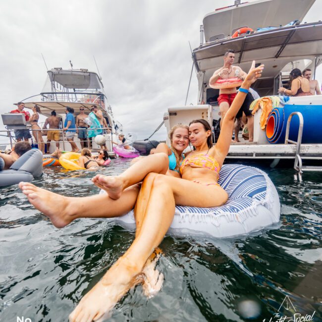 Two women in swimsuits relax on an inflatable raft, smiling and posing for the camera. Behind them, several people enjoy a day on docked yachts, part of The Yacht Social Club's vibrant boat parties in Sydney. The sky is overcast, with various water floaties around.