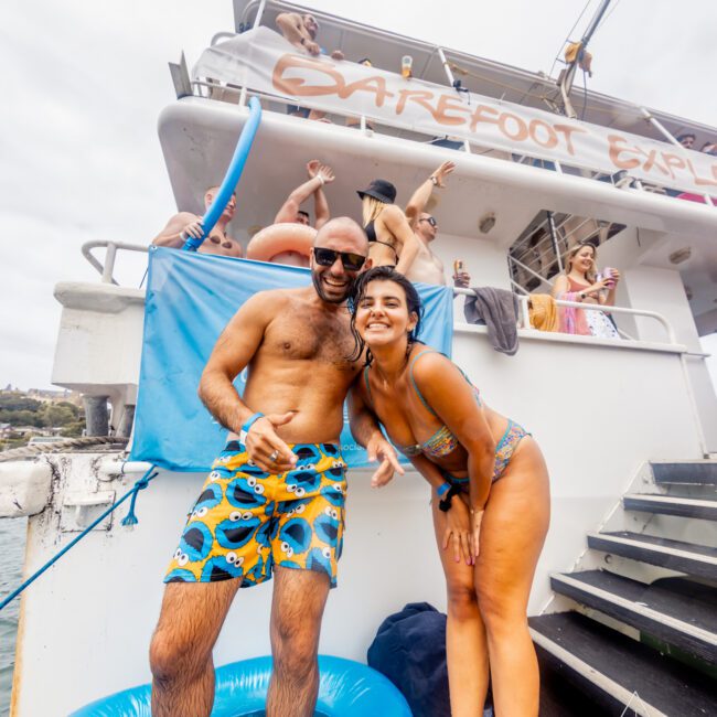 A smiling man and woman in swimsuits pose on the lower deck of a boat during a lively gathering. The man, wearing blue trunks, is holding a drink. Several other partygoers are visible on the upper deck, enjoying the festive atmosphere. Text reads "No swipes" and "The Yacht Social Club Event Boat Charters.