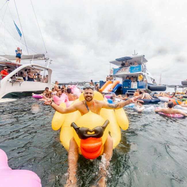 A man smiles and spreads his arms wide while sitting on a large yellow rubber duck float in the water. He is surrounded by other people on various inflatables, near boats in the background under a cloudy sky. Text in the corner reads "Yacht Social Club," showcasing the lively atmosphere of The Yacht Social Club Event.