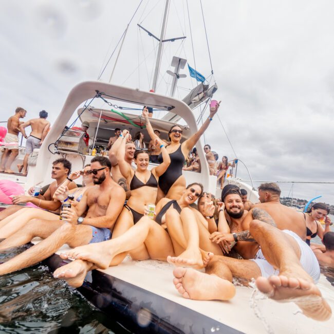 A group of people in swimwear enjoying themselves on a luxury yacht. Some are sitting and lying down while others are standing and cheering, holding drinks. The boat is on the water, and the atmosphere is festive. In the background, more yachts can be seen at The Yacht Social Club Event Boat Charters.