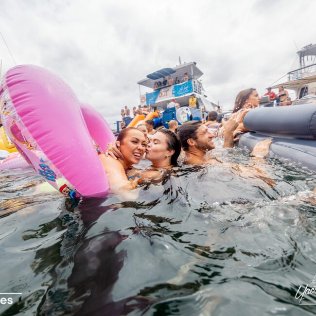 A group of people enjoying a lively pool party in the water, surrounded by inflatable floats and boats. Two women and a man are hugging and smiling while swimming. Other partygoers mingle and have fun against a cloudy sky backdrop, reminiscent of The Yacht Social Club events on Sydney Harbour.