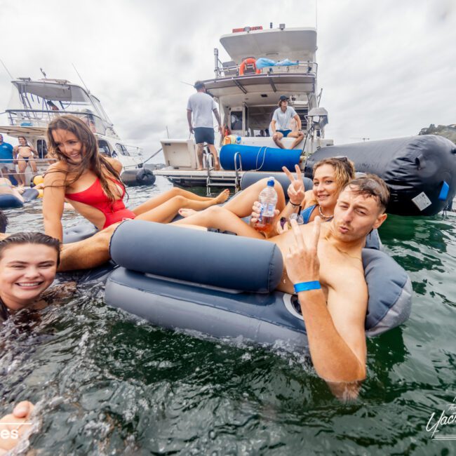 A group of young adults enjoy a day out on the water, lounging on inflatable floats near a boat. One woman in a red swimsuit poses for the camera, while three others make peace signs and smile. More people relax on the boat in the background on a cloudy day, courtesy of Luxury Yacht Rentals Sydney.