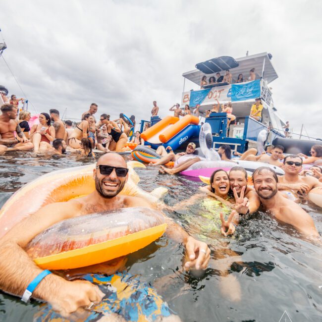 A lively group of people enjoy a yacht party; some are swimming and floating on inflatable toys, while others are on the double-decker yacht in the background. The mood is jubilant under an overcast sky. The scene is branded with "Boat Parties Sydney The Yacht Social Club.