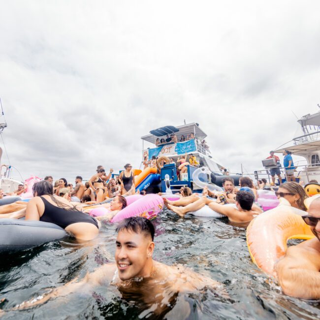 A lively crowd of people are in the water, floating on colorful inflatables and engaging in a festive atmosphere. Boats from The Yacht Social Club are gathered around, with more partygoers seen on the vessels, creating a vibrant and energetic scene under a cloudy sky.