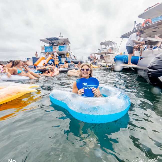 A person in a blue inflatable ring floats in a lively water scene with several boats and people in the background. The person is smiling and holding a circular sign that reads "No swipes." The atmosphere at *The Yacht Social Club Event* is fun and social, with a cloudy sky overhead.