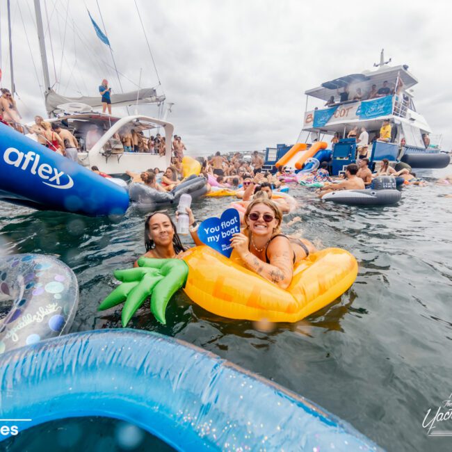 A crowded boat party on a lake with many people floating on inflatable tubes of various shapes and colors. Two women in the foreground are smiling and holding a sign that says "You float my boat." A yacht from The Yacht Social Club Sydney Boat Hire is visible in the background. The sky is overcast.