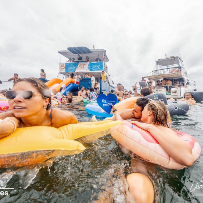 A lively outdoor scene shows a group of young adults enjoying a boat party organized by The Yacht Social Club. Many are floating on inflatable pool toys in the water, soaking up the sun and having fun. Boats with banners and people in swimsuits add to the festive atmosphere on Sydney Harbour.