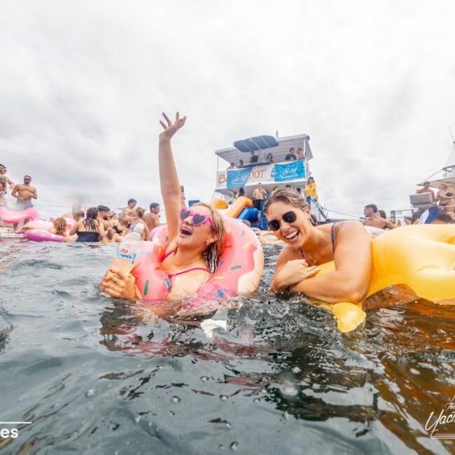 Two women enjoy a lively boat party, smiling and floating on inflatable pool toys in the water. One holds a snack and raises her arm in the air, while the other laughs nearby. Boats and other people from The Yacht Social Club Sydney Boat Hire can be seen in the background.