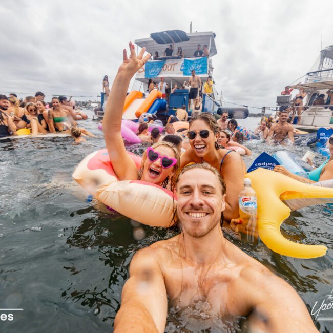 A large group of people, some on inflatables, enjoy a lively pool party near a yacht. A man in the foreground takes a selfie with two women, all smiling and laughing. The background features guests from The Yacht Social Club Sydney Boat Hire and two yachts against a cloudy sky.