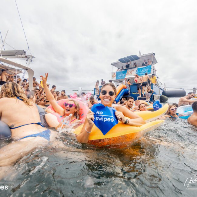 People having fun at a boat party, many in swimsuits and on inflatables in the water. In the foreground is a person riding a banana-shaped float, holding a blue flag that says "No swipes." A large boat with more people partying is visible in the background. Discover Boat Parties Sydney The Yacht Social Club.