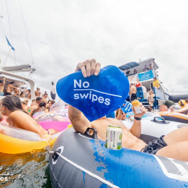 A person lounging on an inflatable raft holds up a blue sign that reads "No swipes" amid a floating party near a boat. Surrounding them are other partygoers on inflatables, enjoying drinks and the water, epitomizing the vibrant atmosphere of The Yacht Social Club Sydney Boat Hire.