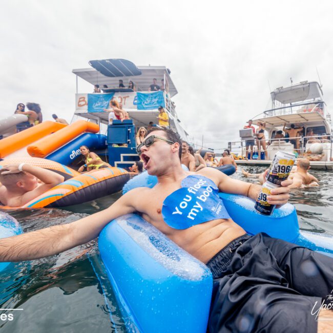 A man relaxes on a blue inflatable raft in a body of water, holding a can and wearing sunglasses. People are enjoying themselves around him on other inflatables and in the water. Two large boats can be seen in the background. Text reads "Yacht Social Club." Enjoy Boat Parties Sydney The Yacht Social Club!